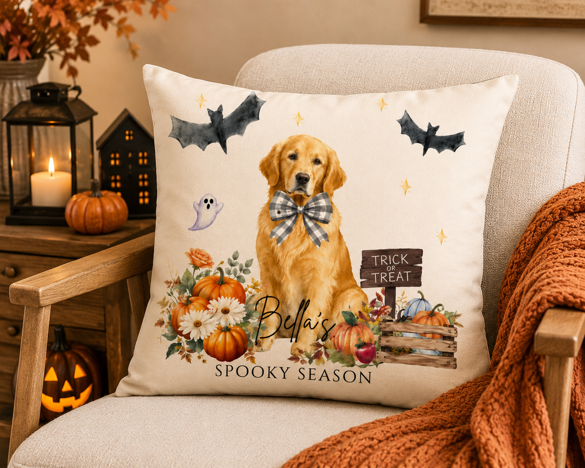 A square decorative throw pillow displayed on an upholstered armchair in a cozy autumn room. The pillow features a printed illustration of a golden retriever wearing a plaid bow, surrounded by pumpkins, flowers, bats, a ghost, and the text 'Bella's Spooky Season.