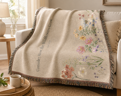 A beige woven throw blanket draped over a white sofa in a bright living room. The blanket features a floral print with pink, yellow, blue, and white wildflowers, vertical text reading 'Conside How the Wildflowers Grow', and fringed edges.