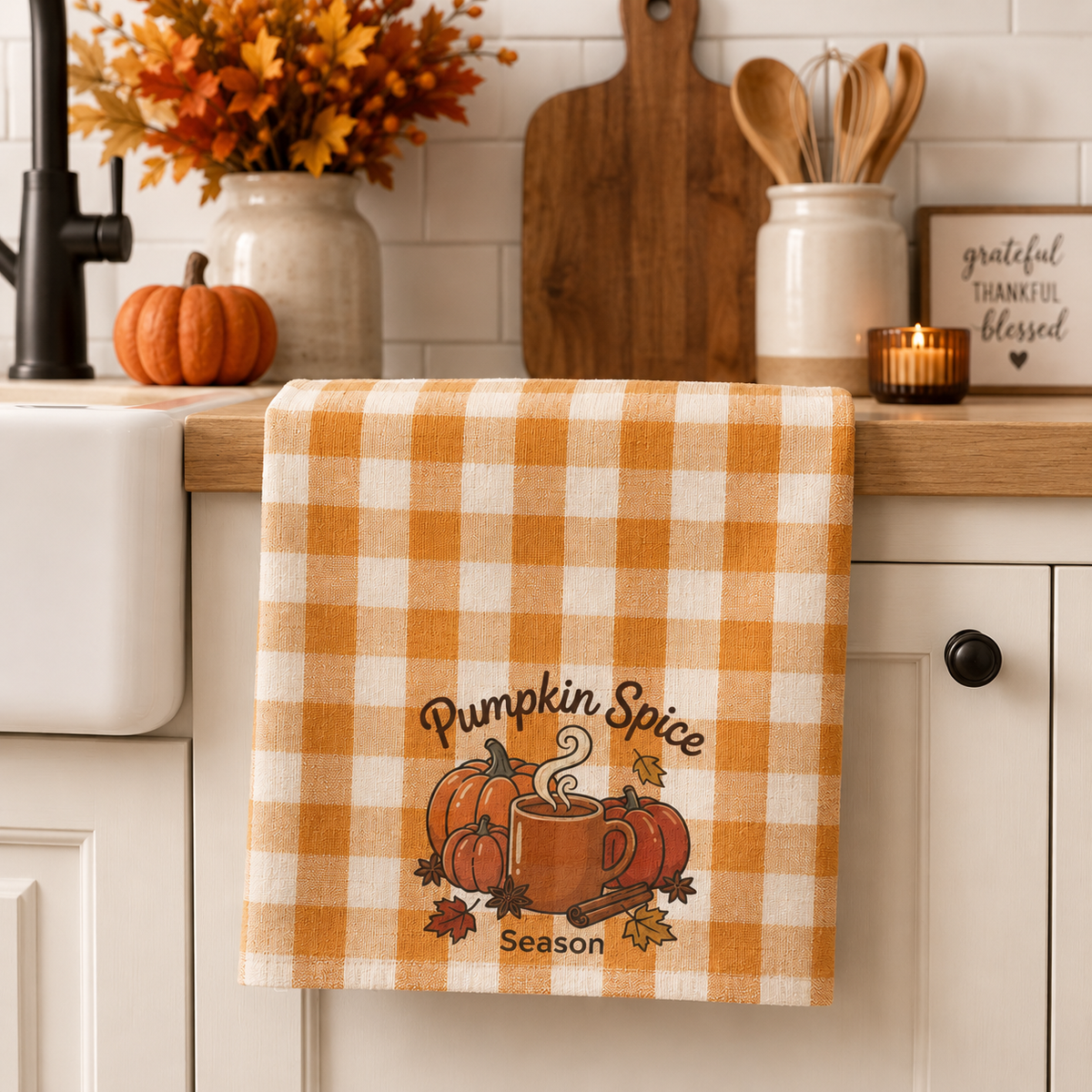 A kitchen towel with an orange and cream gingham pattern hangs over a white kitchen sink. The towel features a 'Pumpkin Spice Season' graphic with pumpkins, a mug, and autumn leaves.