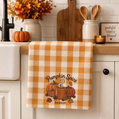 A kitchen towel with an orange and cream gingham pattern hangs over a white kitchen sink. The towel features a 'Pumpkin Spice Season' graphic with pumpkins, a mug, and autumn leaves.