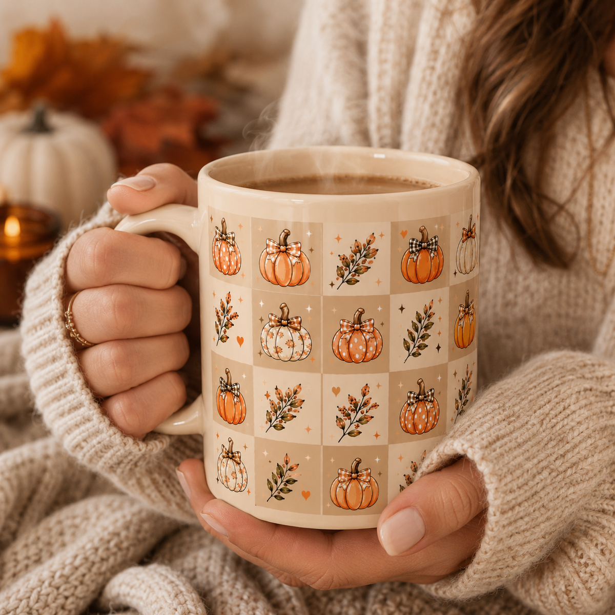 A person in a cozy cream knit sweater holds a ceramic mug decorated with a grid of orange pumpkins and botanical motifs on a warm beige background. The mug is filled with a hot beverage, and the scene is softly lit with autumn decor blurred in the background.