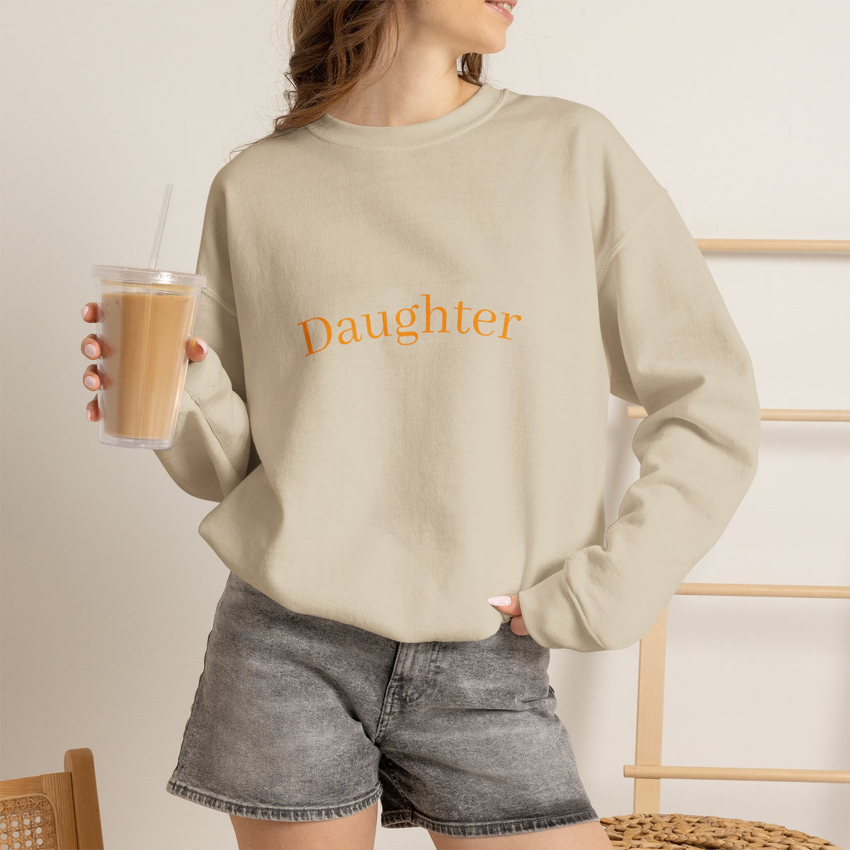 Person wearing a beige sweatshirt with a logo, holding a drink, on a neutral background