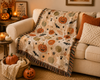 A cozy beige throw blanket with a Halloween print of pumpkins, bats, ghosts, and stars is draped over a cream-colored sofa in a warmly lit living room. The blanket features a fringed edge and is styled with seasonal props including pumpkins, candles, and pillows.