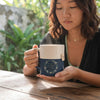 A woman holding a ceramic mug with a cream-colored top half and a navy blue bottom half with a floral wreath design and cursive text. The setting is outdoors on a wooden table with a blurred green garden background.