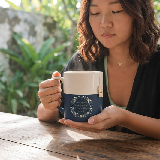 A woman holding a ceramic mug with a cream-colored top half and a navy blue bottom half with a floral wreath design and cursive text. The setting is outdoors on a wooden table with a blurred green garden background.